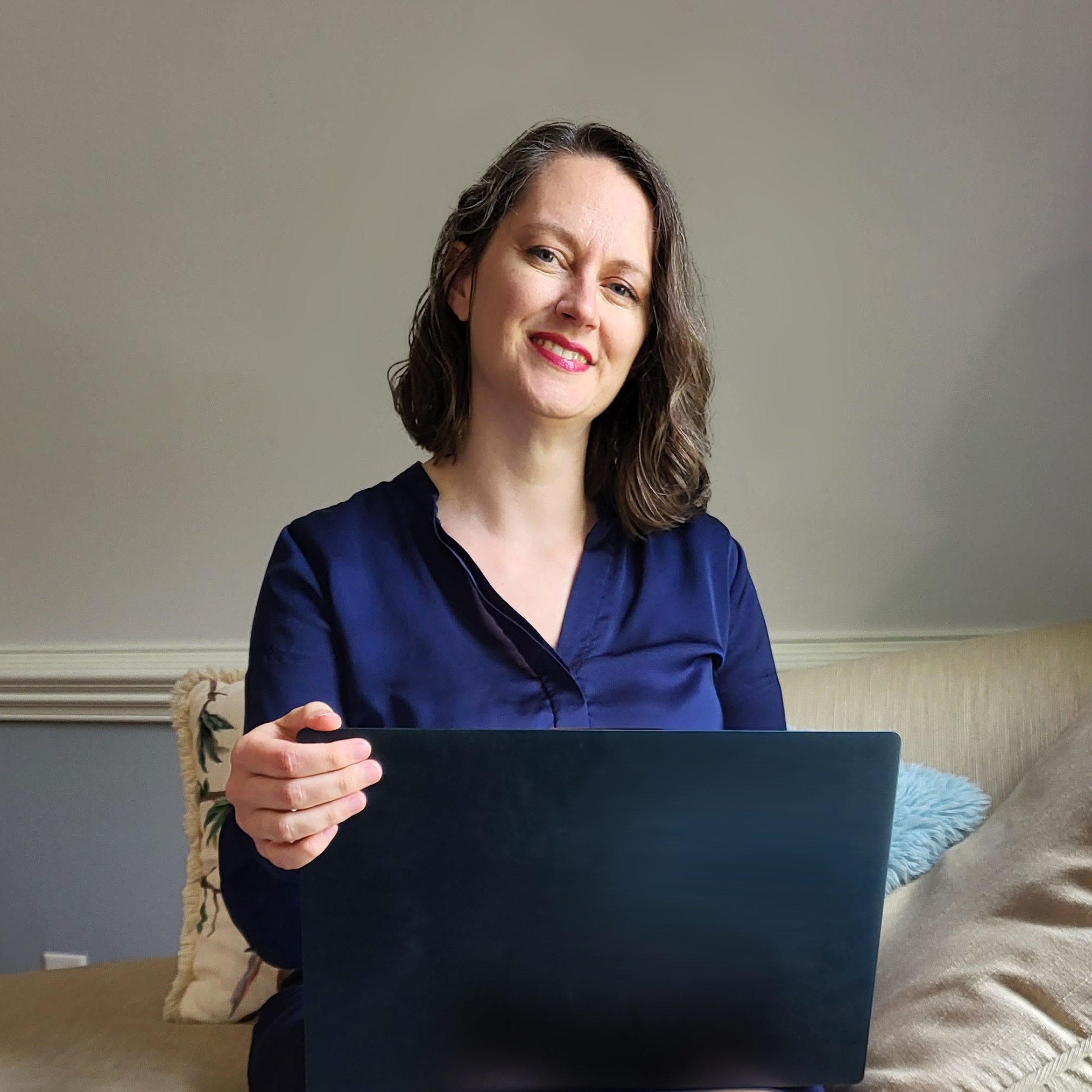 Jordan McCollum, a white woman in her forties with shoulder-length brown wavy hair, in a navy shirt, sitting on a couch holding a laptop