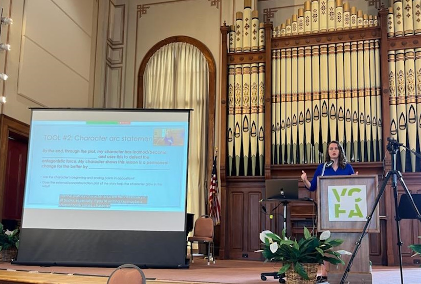 Jordan McCollum, a white woman in her forties with long brown hair, wearing a blue shirt, teaching in a chapel in front of an ornate set of organ pipes. A screen with information about character arcs is on the left.